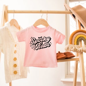May include: Pink baby t-shirt with black text that says "Spooky Babe" with a spiderweb and bat graphic.