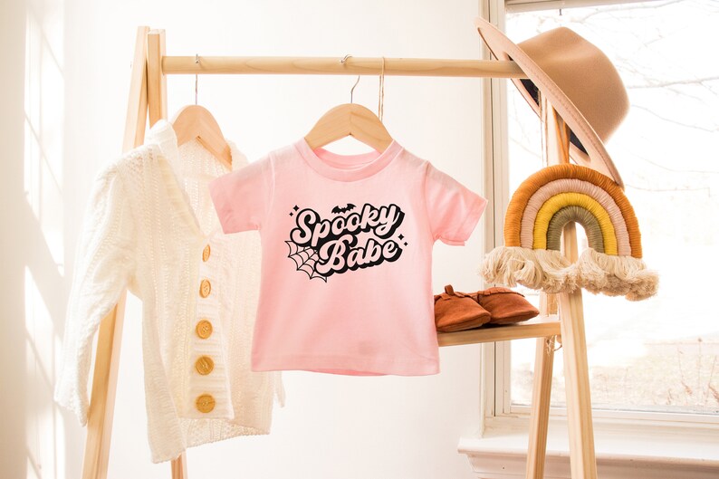 May include: Pink baby t-shirt with black text that says "Spooky Babe" with a spiderweb and bat graphic.
