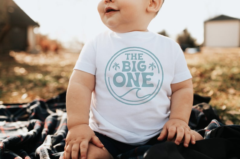 May include: A white baby onesie with a blue circle graphic that says "The Big One" with palm trees and a wave.
