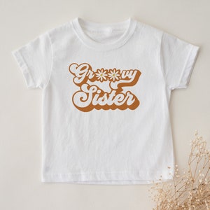 May include: White t-shirt with a brown retro-style graphic that says "Groovy Sister" with a daisy in the middle of the "o".