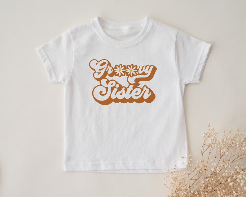 May include: White t-shirt with a brown retro-style graphic that says "Groovy Sister" with a daisy in the middle of the "o".