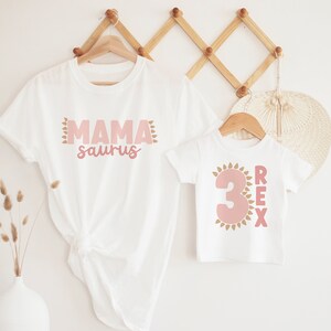May include: Two white t-shirts with pink and gold dinosaur-themed designs. The larger shirt says "Mama saurus" and the smaller shirt says "3 REX".