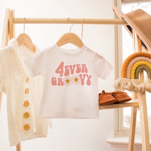 K&ouml;nnte beinhalten: Ein wei&szlig;es T-Shirt mit dem Text "4EVER GROOVY" in Rosa, mit G&auml;nsebl&uuml;mchen-Akzenten. Eine cremefarbene Strickjacke und ein brauner Hut sind ebenfalls zu sehen. Die Artikel werden auf einem Holzst&auml;nder mit einem Wandbehang in Regenbogenfarben pr&auml;sentiert.