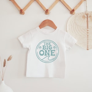 May include: White t-shirt with a blue circle graphic that says "THE BIG ONE" with palm trees on either side of the word "BIG".