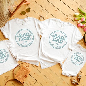 May include: Four white t-shirts with blue text and graphics. The shirts say "Rad Mom", "Rad Dad", "Rad Bro", and "The Big One".