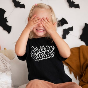 May include: A young child wearing a black t-shirt with the text "Spooky Babe" in white. The shirt has a spiderweb and bat graphic. The child is covering their eyes with their hands and smiling.