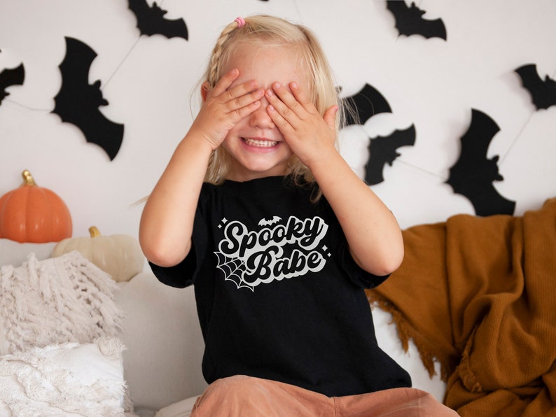 May include: A young child wearing a black t-shirt with the text "Spooky Babe" in white. The shirt has a spiderweb and bat graphic. The child is covering their eyes with their hands and smiling.