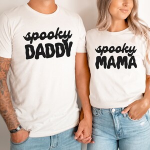 May include: A couple wearing white t-shirts with the text "spooky daddy" and "spooky mama" in black. The man is wearing blue jeans and has tattoos on his arm. The woman is wearing blue jeans and has her hair down.
