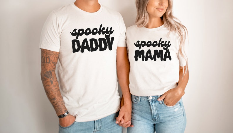 May include: A couple wearing white t-shirts with the text "spooky daddy" and "spooky mama" in black. The man is wearing blue jeans and has tattoos on his arm. The woman is wearing blue jeans and has her hair down.