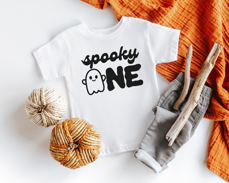 May include: White baby onesie with black text that reads "spooky one" and a ghost graphic. The onesie is laying on a white surface with a brown woven pumpkin, a white woven pumpkin, and a pair of gray pants.