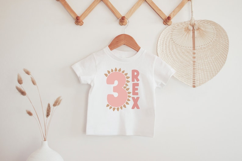 May include: White t-shirt with a pink number 3 surrounded by gold sun rays and the word "REX" in pink letters.