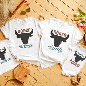 May include: Four white t-shirts with a black bull silhouette and the words "Rodeo" in brown, "mama", "daddy", "brother", and "my first Rodeo" in teal. The shirts are laid out on a wooden surface with a straw hat, flowers, and a woven bag.