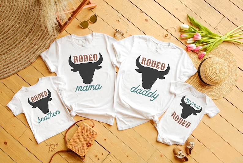 May include: Four white t-shirts with a black bull silhouette and the words "Rodeo" in brown, "mama", "daddy", "brother", and "my first Rodeo" in teal. The shirts are laid out on a wooden surface with a straw hat, flowers, and a woven bag.