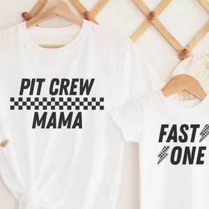May include: Two white t-shirts with black text and checkered racing flag designs. The larger shirt says "Pit Crew Mama" and the smaller shirt says "Fast One" with a lightning bolt.