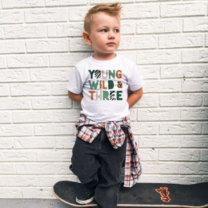 May include: A young boy wearing a white t-shirt with the text "YOUNG WILD & THREE" in green, brown, and orange with animal print patterns. He is standing on a skateboard with his hands in his pockets.