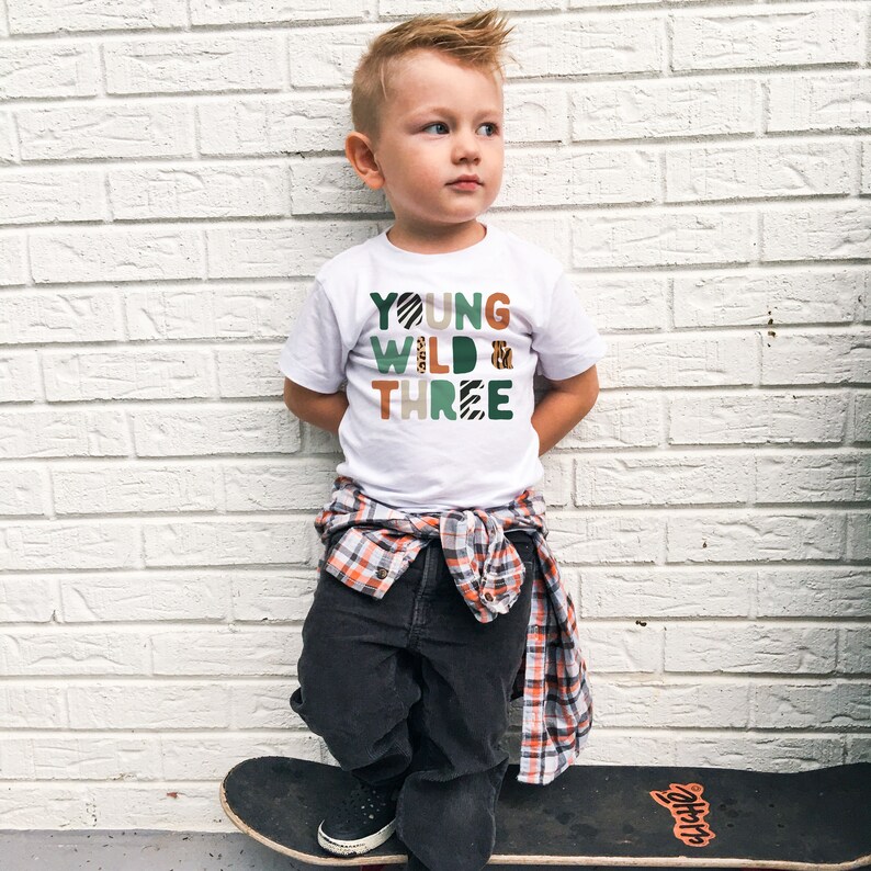May include: A young boy wearing a white t-shirt with the text "YOUNG WILD & THREE" in green, brown, and orange with animal print patterns. He is standing on a skateboard with his hands in his pockets.