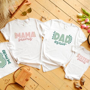 May include: White t-shirts with a dinosaur theme. The shirts say "Mama Saurus", "Dad Osaur", "Sister Saurus", and "Rex 3".
