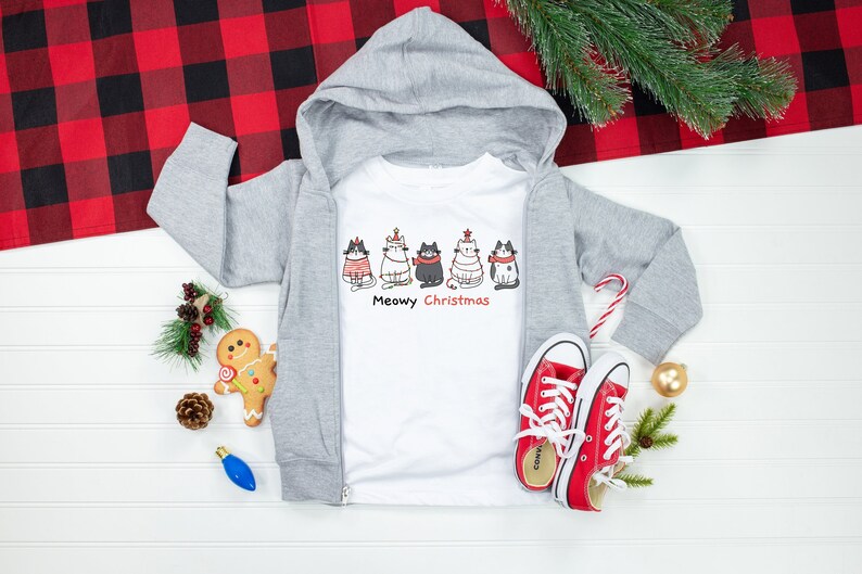 May include: A white hooded sweatshirt with a zipper and a white t-shirt underneath. The t-shirt has a graphic of five cats wearing Christmas hats and the text "Meowy Christmas".