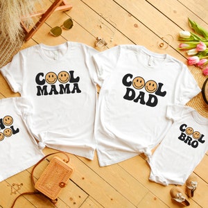 May include: Four white t-shirts with a retro design featuring smiling faces and the text "Cool Mama", "Cool Dad", "Two Cool", and "Cool Bro".