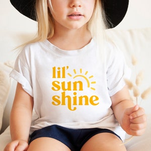 May include: A white t-shirt with a yellow graphic that says "lil' sun shine" and a stylized sun graphic.