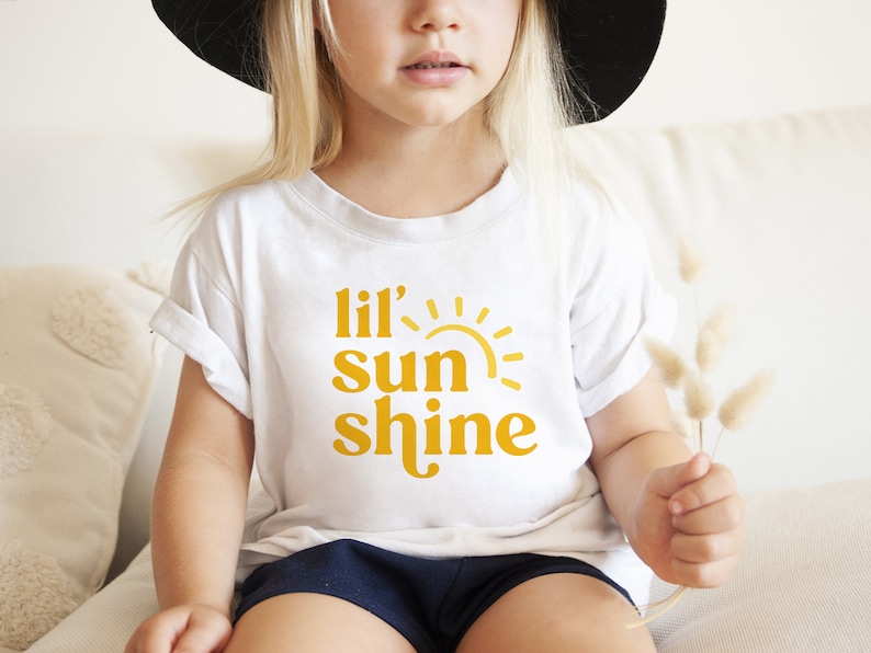May include: A white t-shirt with a yellow graphic that says "lil' sun shine" and a stylized sun graphic.