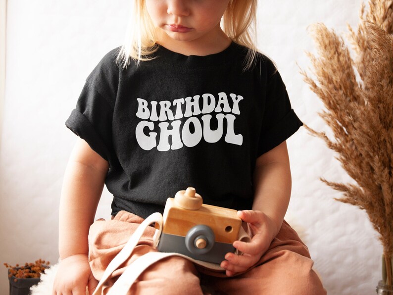 May include: A child wearing a black t-shirt with the text "Birthday Ghoul" in white retro-style lettering. The child is holding a wooden toy camera.