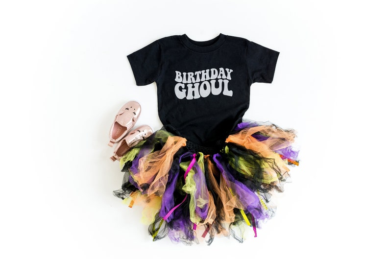 May include: Black t-shirt with the text "Birthday Ghoul" in white, a multi-colored tulle skirt, and pink shoes. Halloween costume for a child.