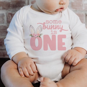 May include: A white long-sleeved onesie with pink bunny ears and a pink floral crown. The onesie has the text "some bunny is one" in pink.