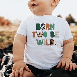 May include: A white t-shirt with the text "Born Two Be Wild." in colorful letters with animal print patterns. The letters are in a variety of colors, including green, brown, orange, and black. The shirt is being worn by a young child.