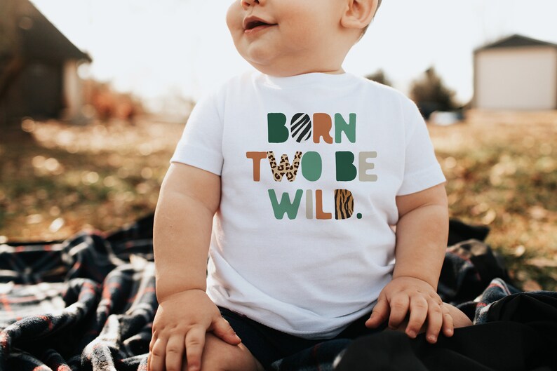 May include: A white t-shirt with the text "Born Two Be Wild." in colorful letters with animal print patterns. The letters are in a variety of colors, including green, brown, orange, and black. The shirt is being worn by a young child.