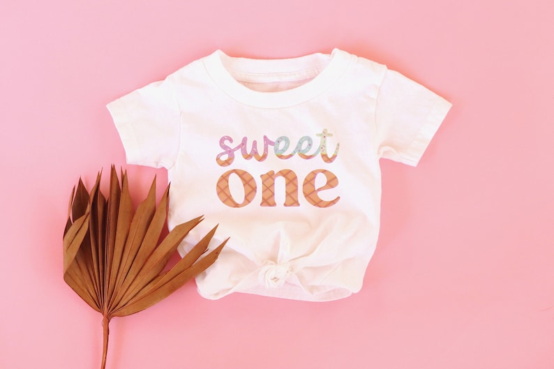 May include: White baby onesie with a pink, blue, and orange "sweet one" graphic.