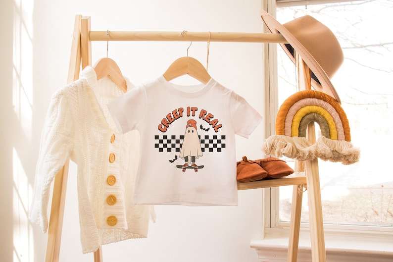 May include: White t-shirt with a graphic of a ghost wearing a red hat and skateboarding. The text "Creep it Real" is printed above the ghost. The t-shirt is hanging on a wooden rack.