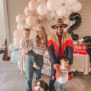 May include: A family of four poses for a photo with a dog. The parents are wearing red and white shirts with the words "Pit Crew" on them. The two children are wearing white shirts with the words "Two Fast" and "Pit Crew" on them. The dog is wearing a red bandana. The background is decorated with balloons and a black and white checkered race track.
