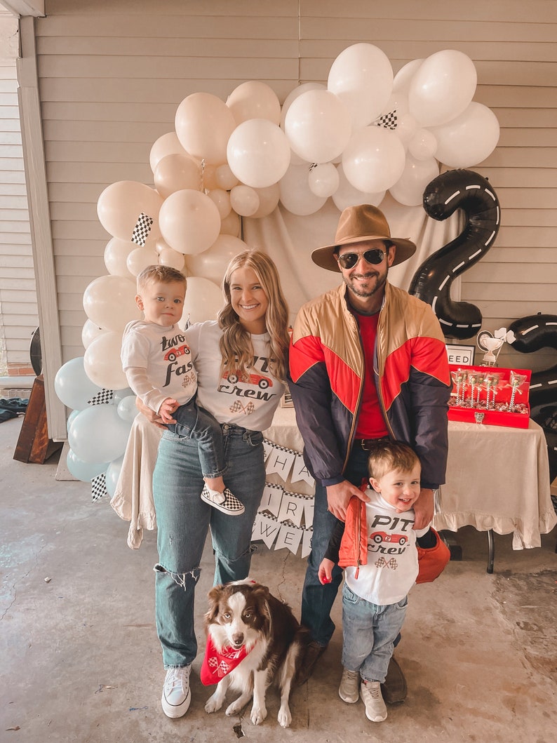 May include: A family of four poses for a photo with a dog. The parents are wearing red and white shirts with the words "Pit Crew" on them. The two children are wearing white shirts with the words "Two Fast" and "Pit Crew" on them. The dog is wearing a red bandana. The background is decorated with balloons and a black and white checkered race track.