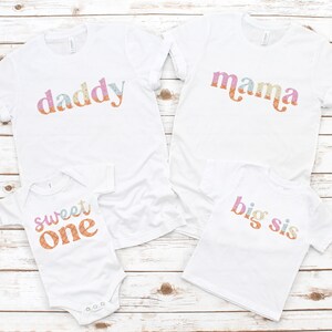 May include: A set of four white t-shirts with colorful text. The shirts say "daddy", "mama", "sweet one", and "big sis".