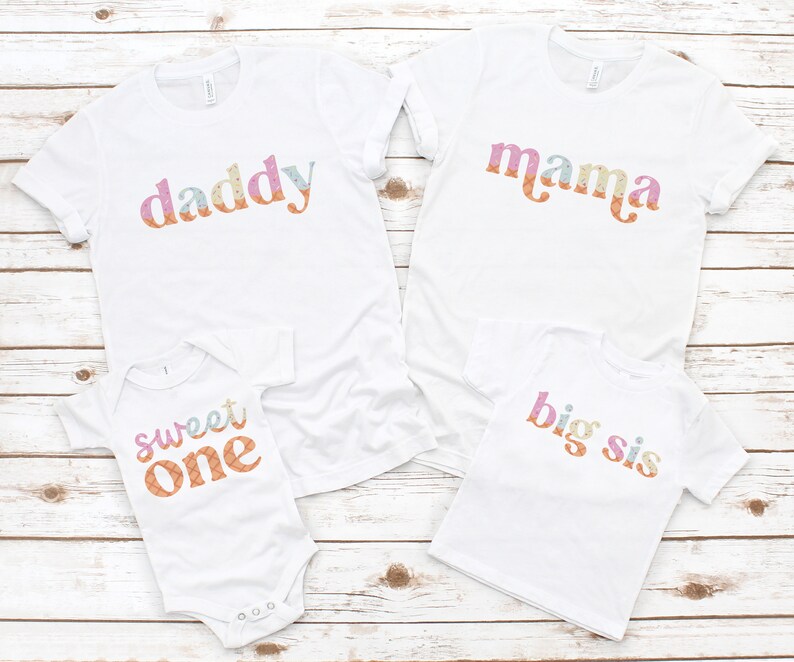 May include: A set of four white t-shirts with colorful text. The shirts say "daddy", "mama", "sweet one", and "big sis".