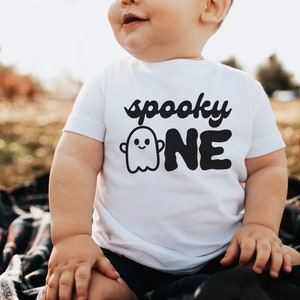 May include: A white onesie with a black ghost and the text "spooky one" on it. A baby is wearing the onesie and sitting on a plaid blanket.