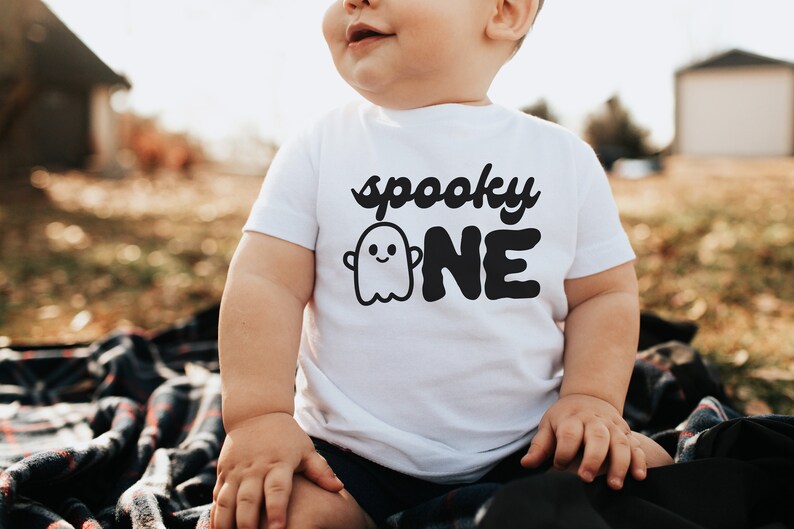 May include: A white onesie with a black ghost and the text "spooky one" on it. A baby is wearing the onesie and sitting on a plaid blanket.