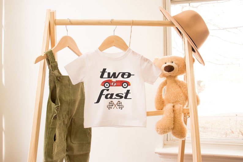 May include: White t-shirt with a red race car graphic and the text "two fast" in black. The shirt is hanging on a wooden rack with a pair of green overalls and a teddy bear.