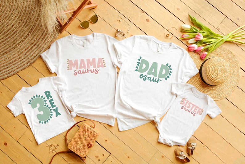 May include: Four white t-shirts with green and pink dinosaur themed text. The shirts say 'Mama saurus', 'Dad osaur', 'Sister saurus', and 'Rex 3'. The shirts are laid out on a wooden surface.