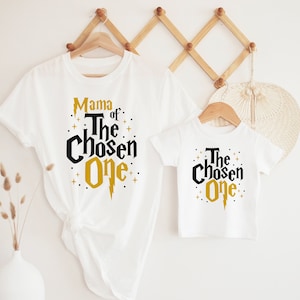 May include: Two white t-shirts with black and gold text. The larger shirt says "Mama of The Chosen One" with a gold lightning bolt. The smaller shirt says "The Chosen One" with a gold lightning bolt.