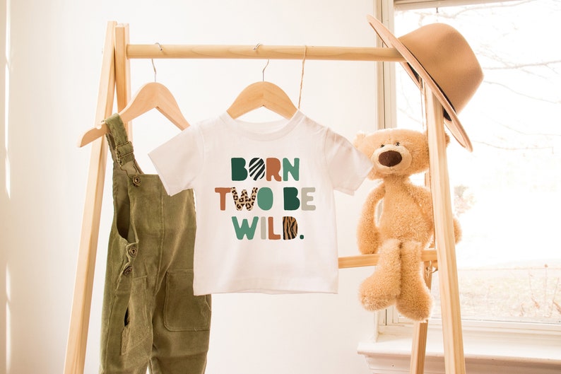 May include: White baby onesie with the text "BORN TWO BE WILD." in colorful letters with animal print patterns. The onesie is hanging on a wooden rack with a stuffed teddy bear and a hat.