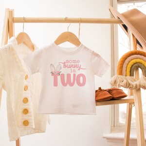 May include: A white baby onesie with a pink bunny graphic and the text "some bunny is two". The onesie is hanging on a wooden rack with a white cardigan and a rainbow macrame hanging on the right.