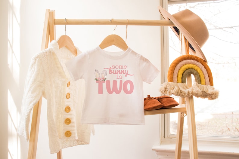May include: A white baby onesie with a pink bunny graphic and the text "some bunny is two". The onesie is hanging on a wooden rack with a white cardigan and a rainbow macrame hanging on the right.