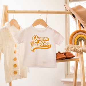 Pu&ograve; includere: Maglietta bianca per neonato con un design retr&ograve; giallo che dice "Queen Bee" con un'illustrazione di un'ape. La maglietta &egrave; appesa a un appendiabiti in legno con un cardigan bianco e un macram&egrave; arcobaleno accanto.