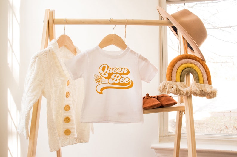Pu&ograve; includere: Maglietta bianca per neonato con un design retr&ograve; giallo che dice "Queen Bee" con un'illustrazione di un'ape. La maglietta &egrave; appesa a un appendiabiti in legno con un cardigan bianco e un macram&egrave; arcobaleno accanto.