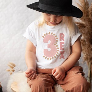 May include: A child wearing a white t-shirt with a pink number "3" inside a brown sun graphic and the word "REX" in brown letters. The child is wearing a black hat and brown pants.