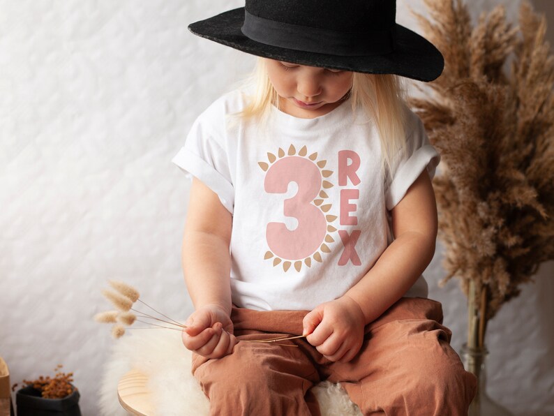 May include: A child wearing a white t-shirt with a pink number "3" inside a brown sun graphic and the word "REX" in brown letters. The child is wearing a black hat and brown pants.