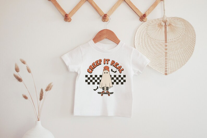May include: White t-shirt with a ghost wearing a red hat and skateboarding. The text "CREEP IT REAL" is above the ghost. The ghost is on a checkered background with bats flying around it.
