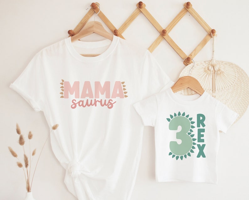 May include: Two white t-shirts with a dinosaur theme. The larger shirt says "Mama saurus" in pink lettering with brown accents. The smaller shirt says "3 Rex" in green lettering with green accents.